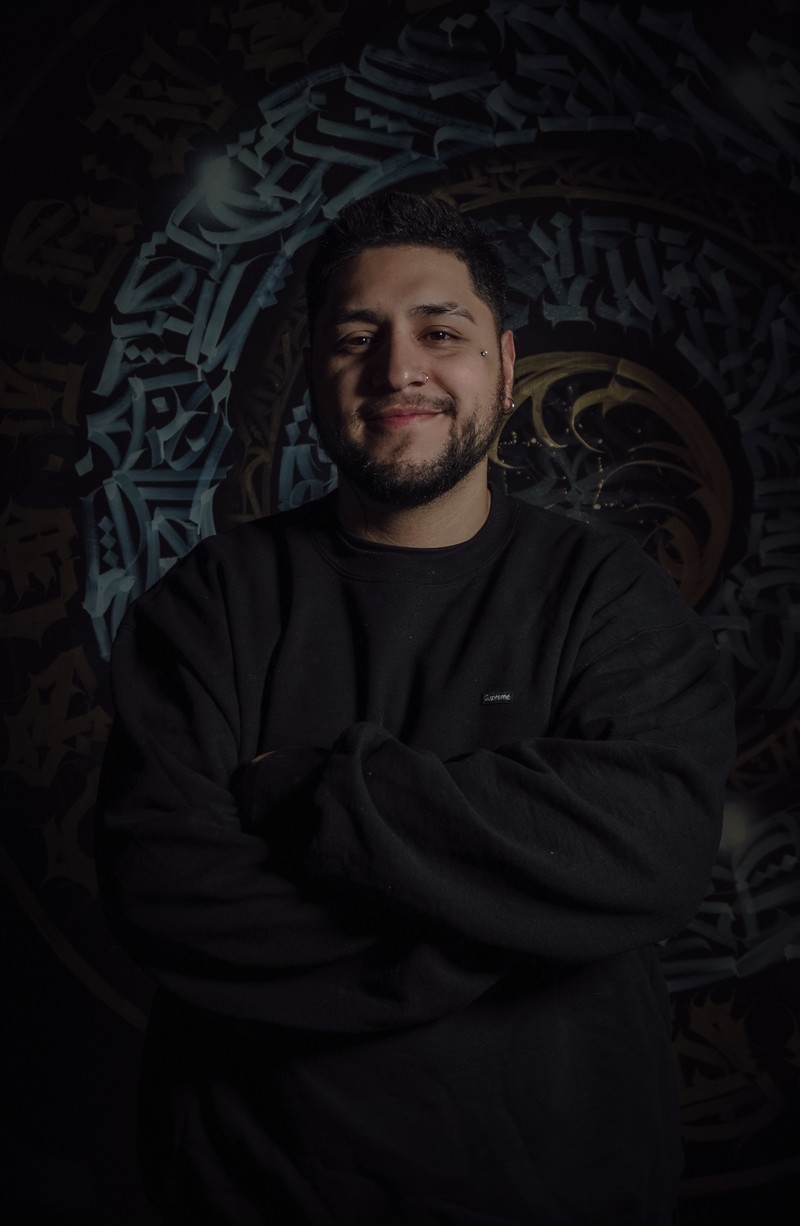Sadone facing the camera with a relaxed expression, wearing black against a calligraphic wall.