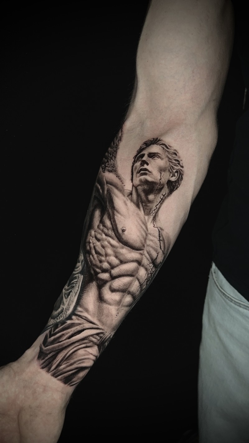 Black and grey forearm tattoo of a sculptural male figure, with modeled anatomy and folds of fabric.
