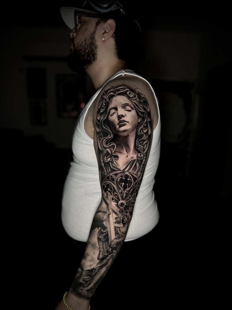 Large black and grey sleeve with a female sculptural figure, ornamental forms and scenes along the arm.